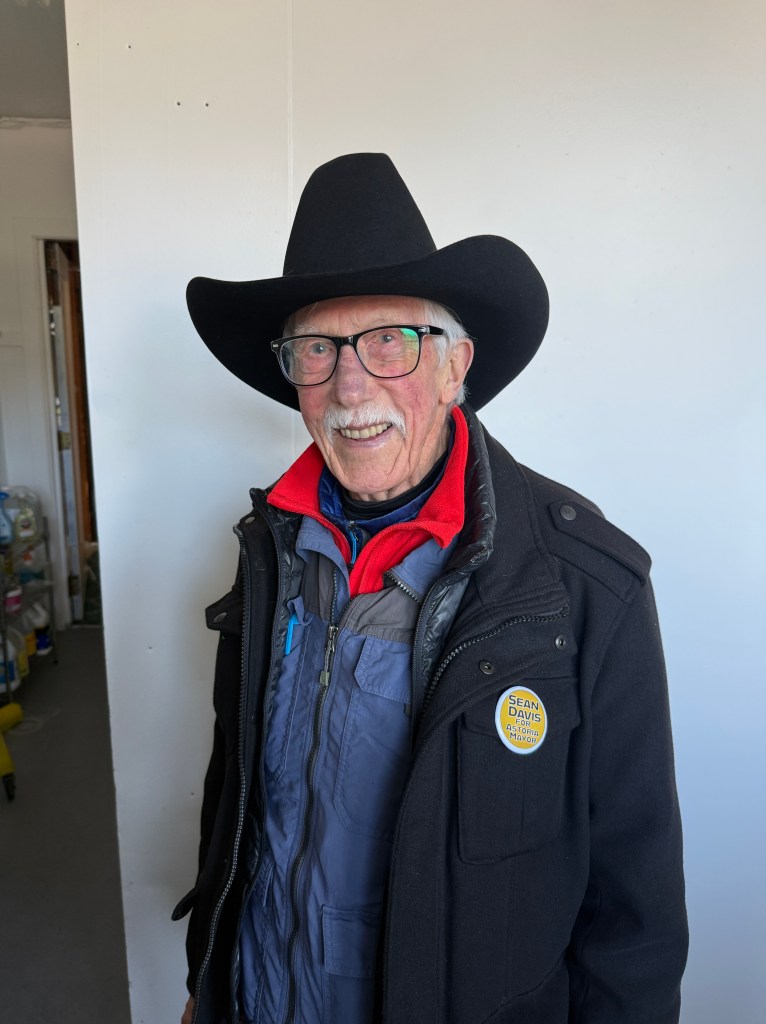 An elderly man wearing a large black cowboy hat, glasses, and a coat with a red collar. He is smiling and stands in front of a plain white wall. A pin on his coat reads 'Sean Davis for Astoria Mayor.'