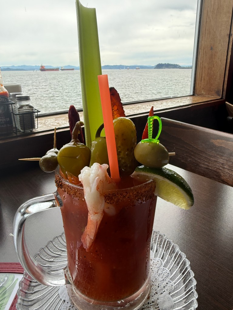 A close-up of a colorful Bloody Mary cocktail garnished with celery, shrimp, olives, pickles, and a lime wedge, sitting on a lace doily with a scenic view of the water in the background.