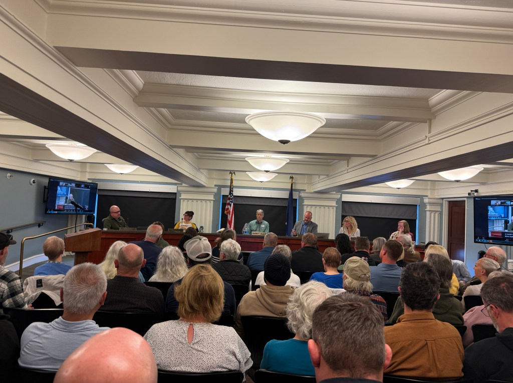 A panel discussion taking place indoors with a diverse audience seated in front of a stage. Panelists, including five individuals, are seated behind a table with microphones, while large screens display visuals. American flags and a professional setting are visible in the background.