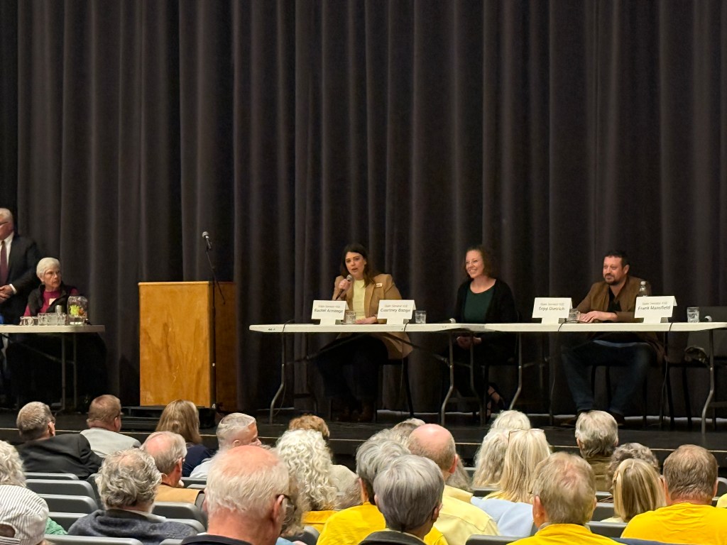 A public forum featuring a panel of speakers at a table, with an audience seated in front. The panel includes four individuals, two women and two men, discussing various topics. A backdrop of curtains is visible behind the speakers.