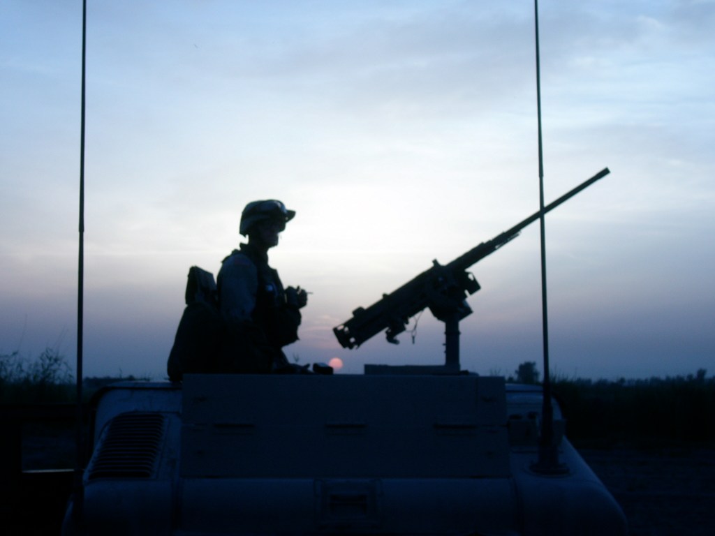 Silhouette of a soldier sitting on a military vehicle, holding a camera, with a mounted machine gun, against a sunset background.