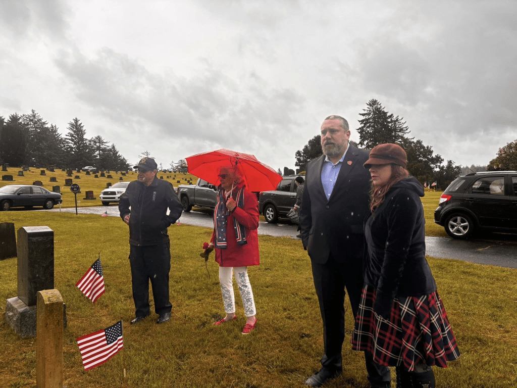 A group of four people standing in a cemetery, with two holding American flags. One person is under a red umbrella, and the weather appears overcast and rainy.