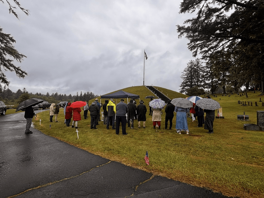 A group of people gathered at a cemetery during a rainy day, some holding umbrellas, near a flag and a tent.
