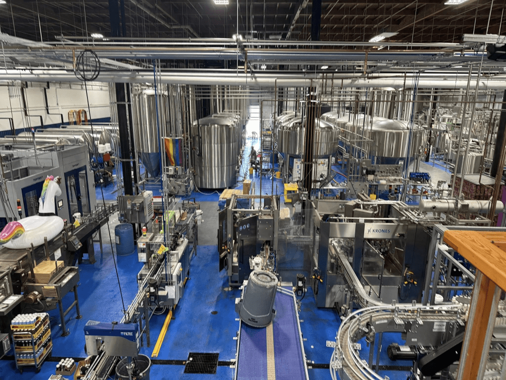 A spacious industrial brewery interior featuring large brewing tanks, machinery, and conveyor belts on a blue floor.