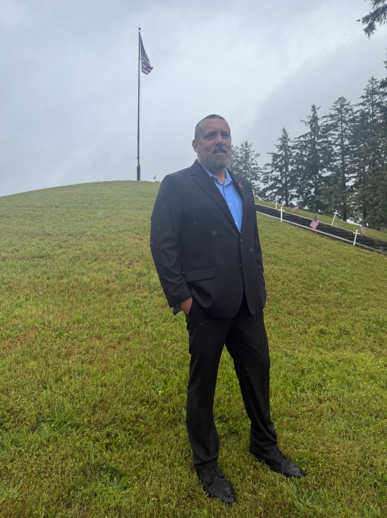 A man in a black suit standing on a grassy hill, with a blue shirt underneath and an American flag in the background. The scene is slightly overcast.