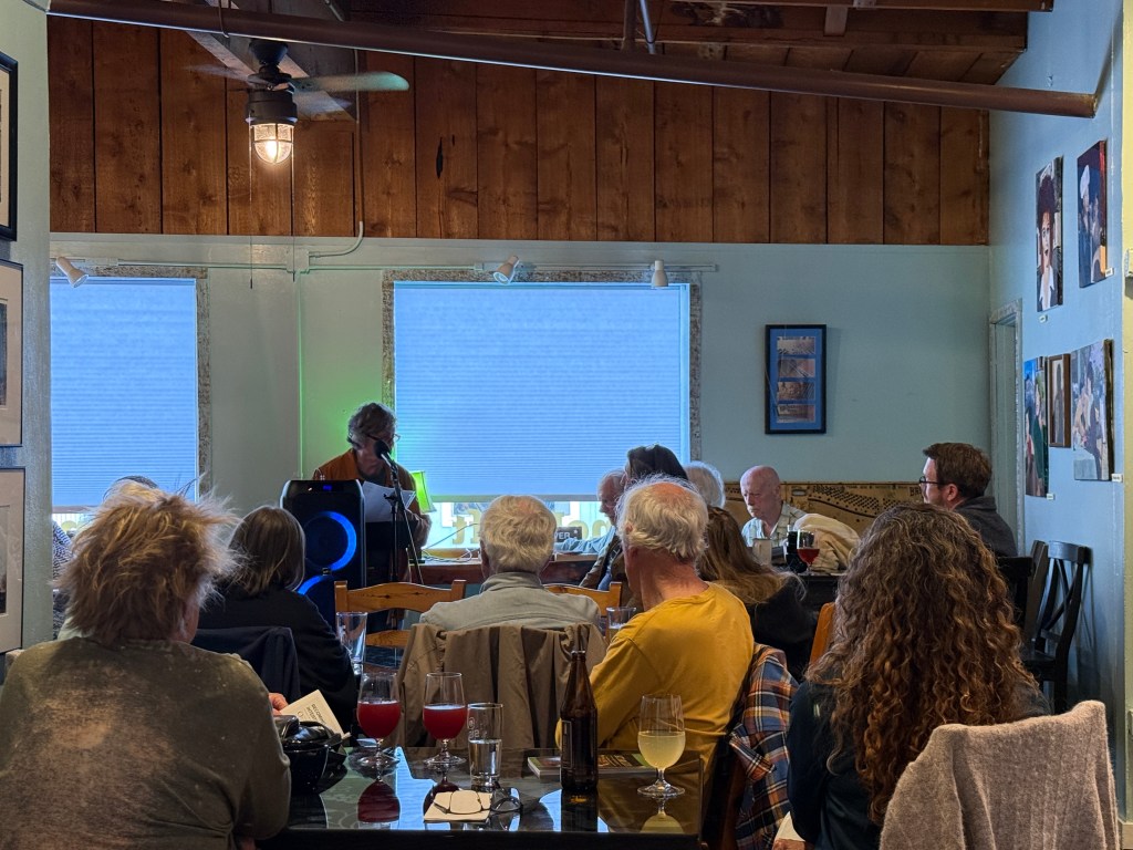 A cozy restaurant setting with a speaker at the front, addressing an audience seated at tables. The room features wooden panels and a few artworks on the walls. Various drinks are visible on the tables.