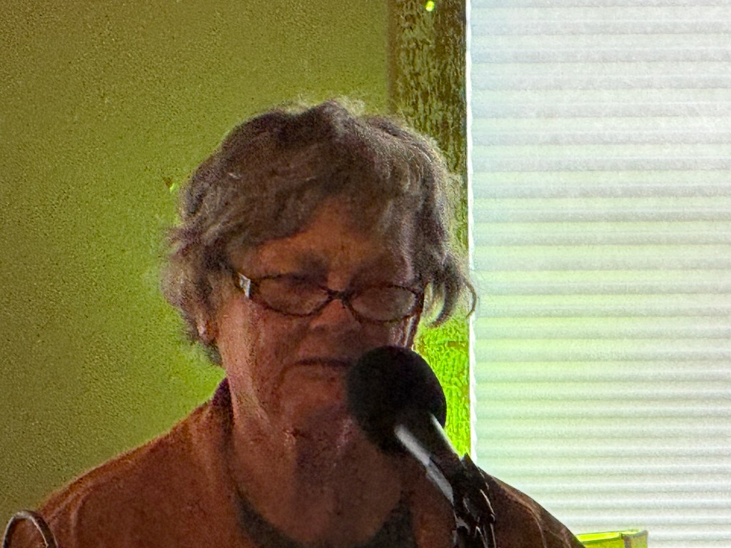 An older woman with glasses speaks into a microphone, set against a background with natural light filtering through a window.