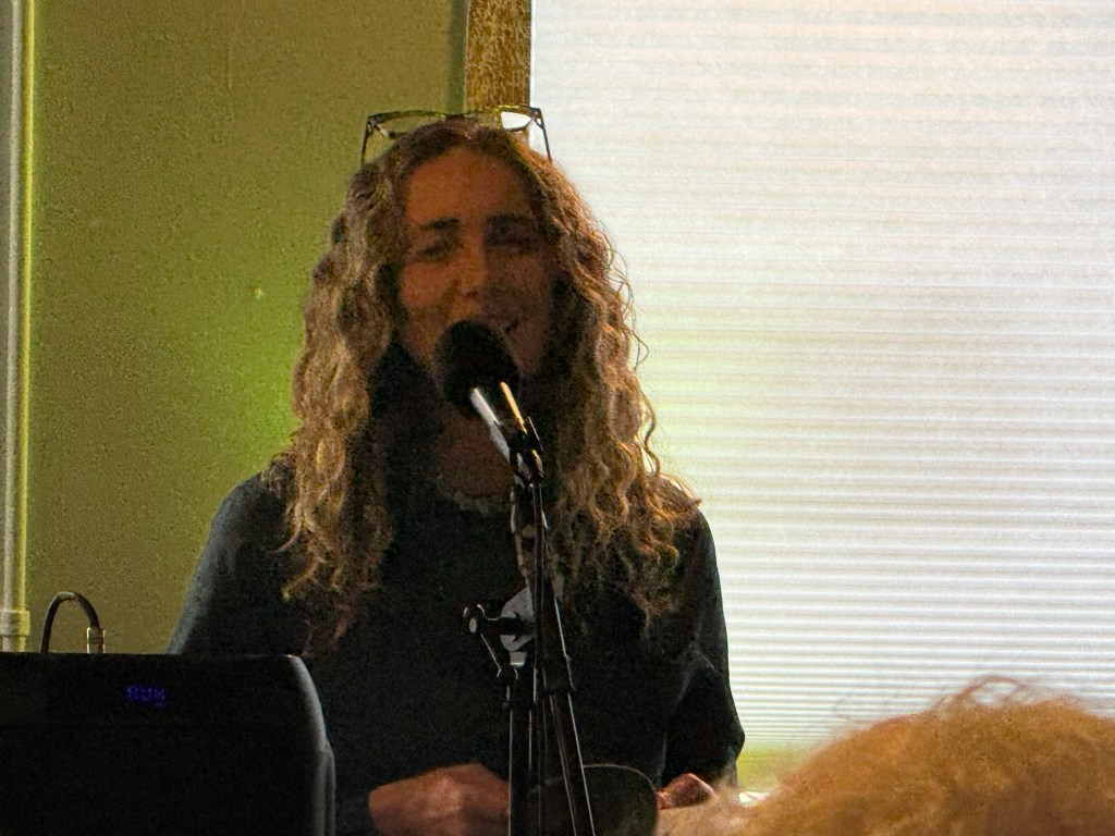 A woman with curly hair singing into a microphone, wearing a dark shirt, in a well-lit indoor setting.