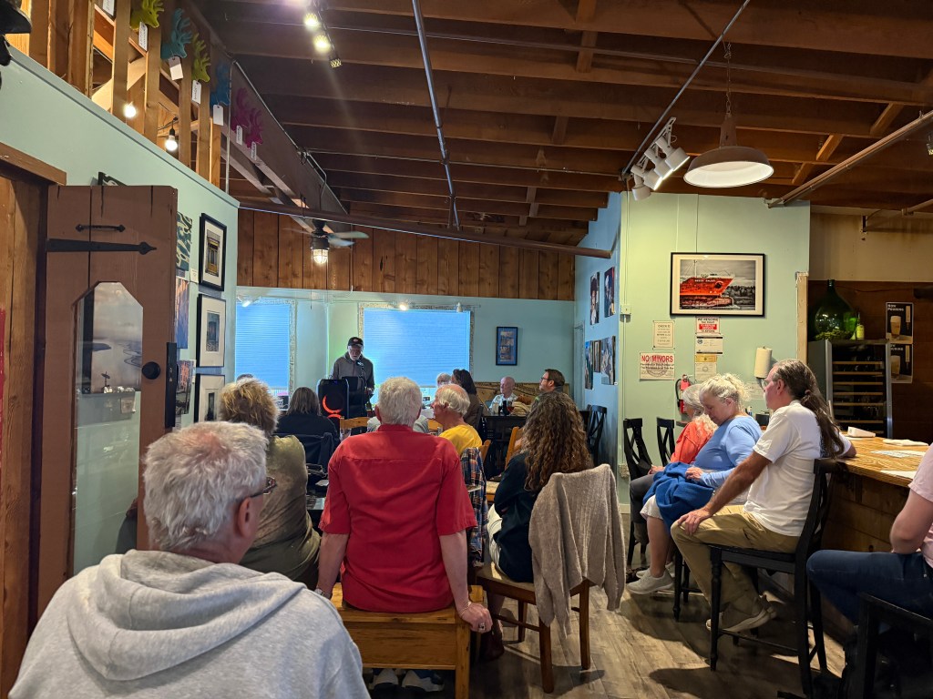 A small crowd gathered in a cozy room for a presentation or performance, with a speaker at the front. The walls are decorated with various framed photographs, and natural light is coming through the windows.