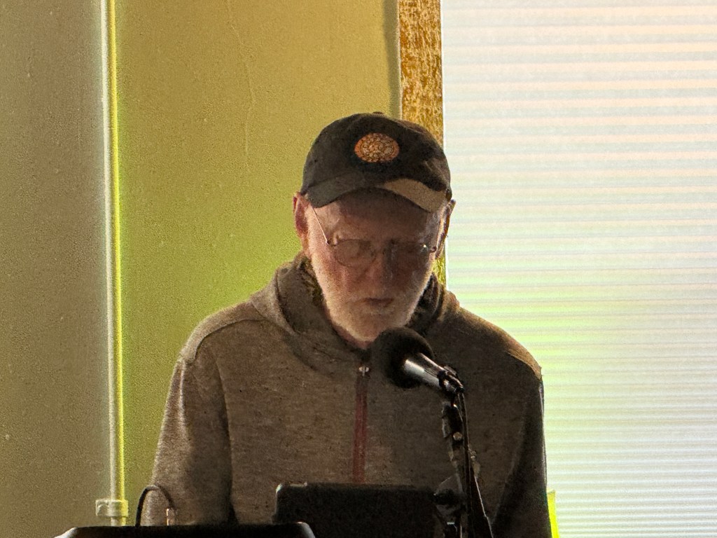 A person with glasses and a cap reading from a podium, illuminated by natural light from a window.