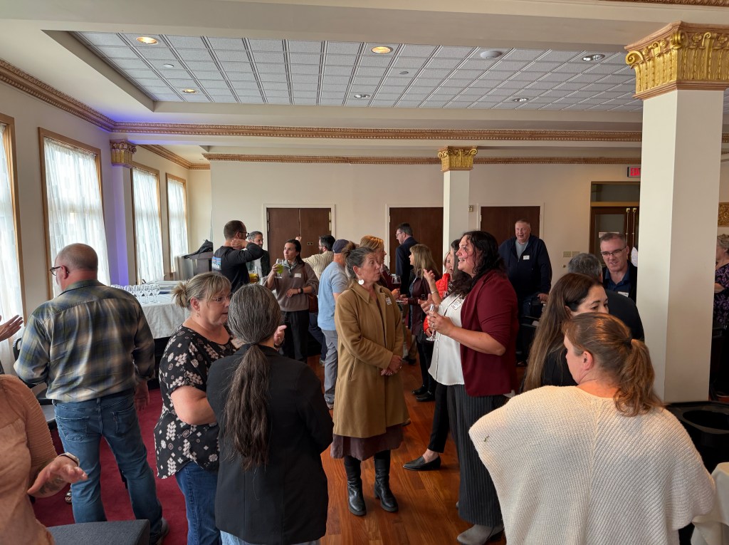 A lively gathering of people in a spacious room, engaging in conversation and socializing. The room features large windows with natural light, elegant decorations, and individuals holding drinks. Attendees are dressed in smart casual attire.