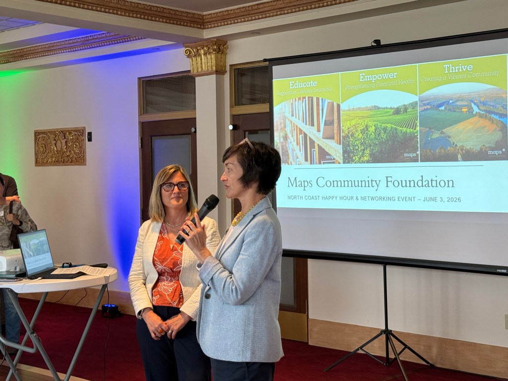 Two women speaking at a community event, with a presentation in the background displaying the Maps Community Foundation logo and themes of education, empowerment, and thriving communities.