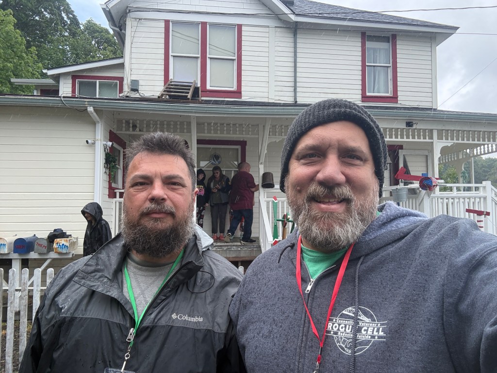 Two men posing for a selfie in front of a white house with red accents, while people are gathered on the porch in the background. The weather appears cloudy or rainy.