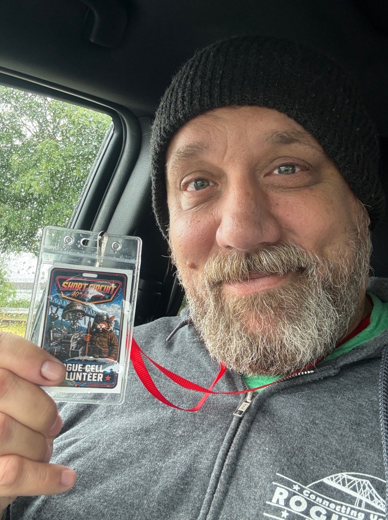 A person wearing a beanie and gray hoodie smiles while holding a volunteer badge in their car. The background shows rain on the window.