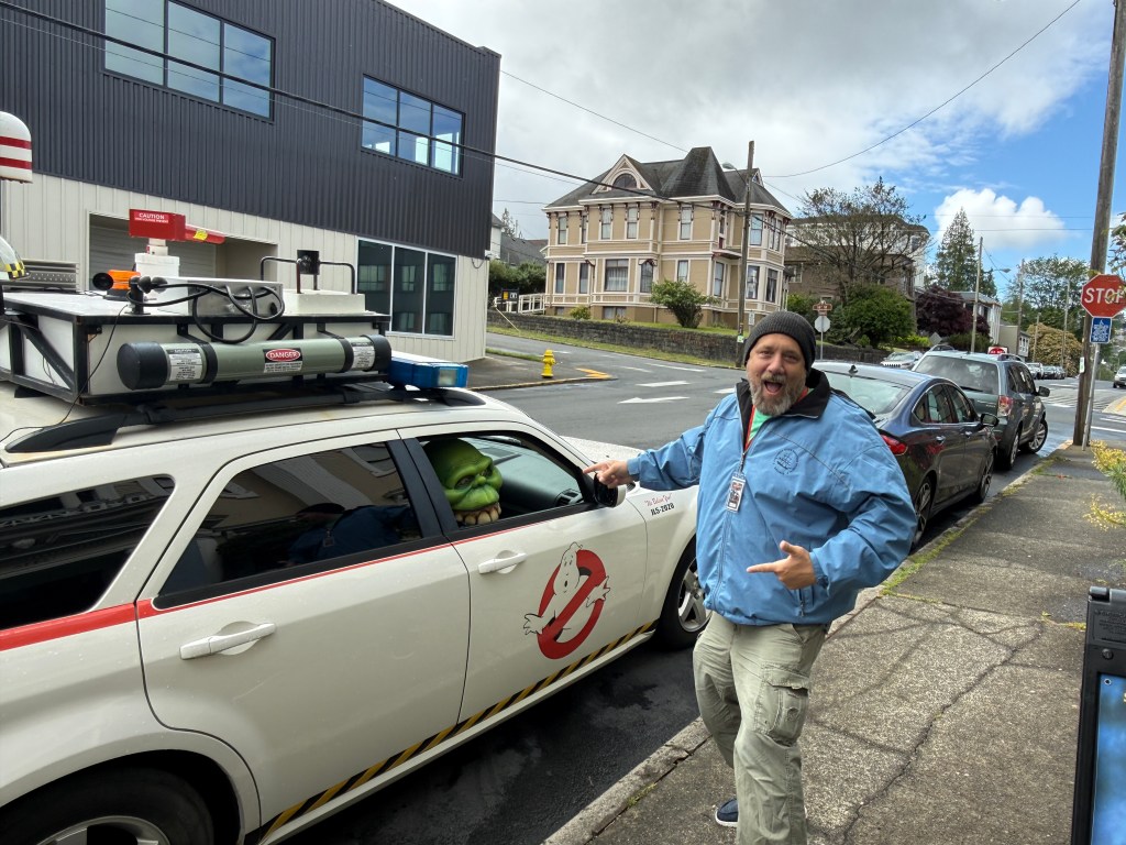 A man in a blue jacket and a knit cap poses excitedly next to a Ghostbusters-themed car, which features a cartoonish green ghost character peeking from the driver's side window. The background includes a residential area with a large house and other parked cars.