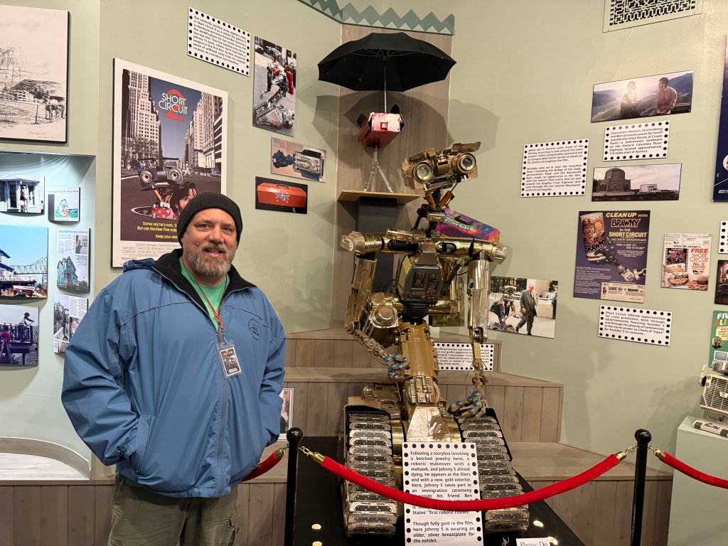 A man with a beard wearing a blue jacket and beanie stands next to a robot statue in a museum setting. The walls are decorated with various movie posters and photographs.