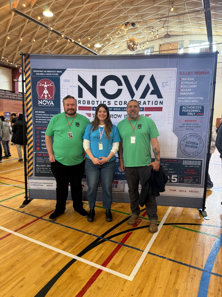 Three people posing in front of a large banner for NOVA Robotics Corporation in a gymnasium. Two individuals are wearing green shirts, and one is in a blue shirt. The banner displays the company's logo and information about their programs.