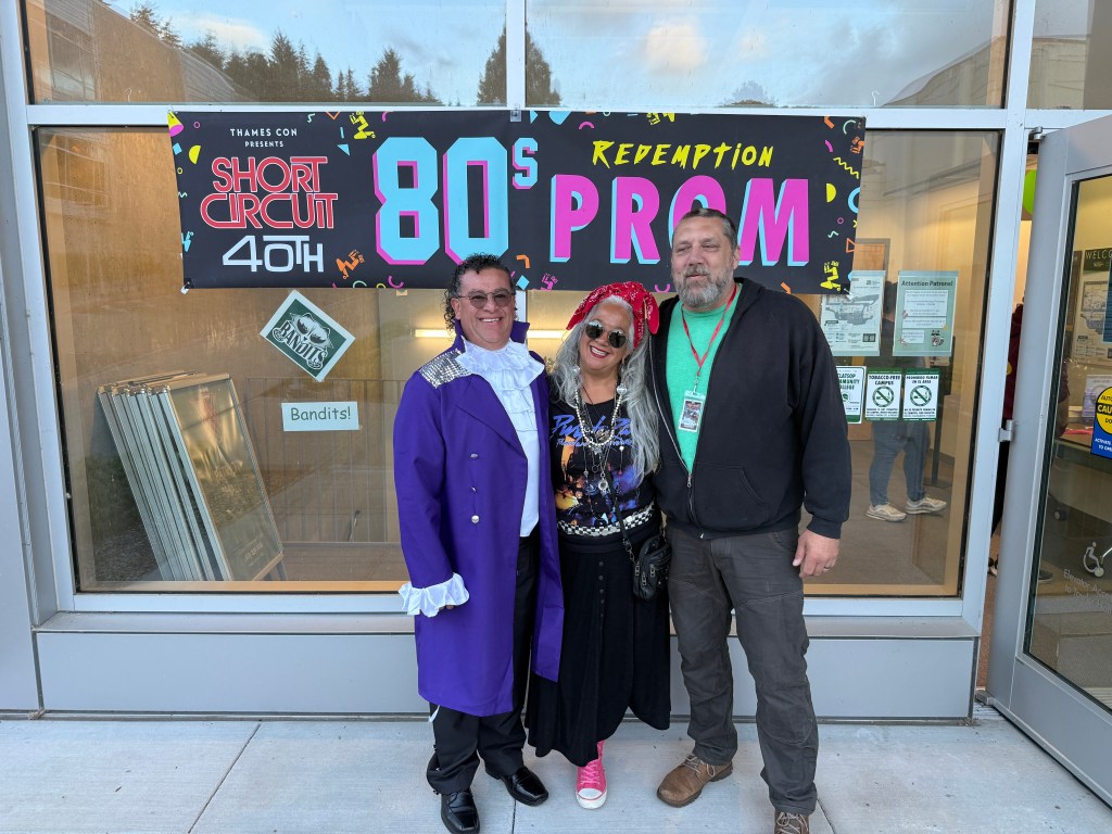 Three individuals pose in front of a window with a colorful banner that reads '80s Redemption Prom' for the 40th anniversary of 'Short Circuit'.