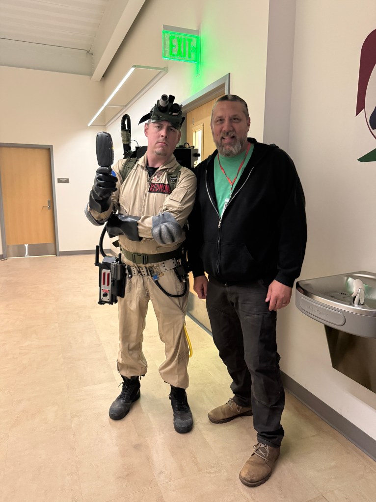 Two men pose for a photo in a hallway. One man is dressed as a Ghostbuster in a beige jumpsuit with a proton pack, while the other man wears casual clothing.