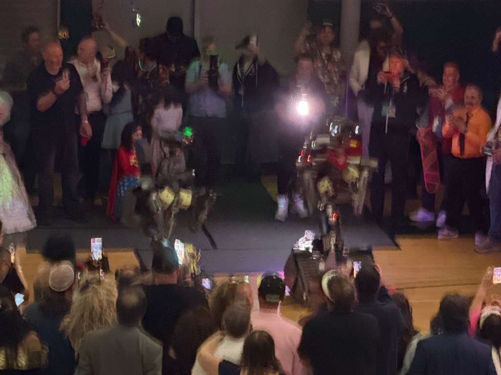 A crowd watches as two robots perform on stage, with people capturing the moment on their phones at a lively event.