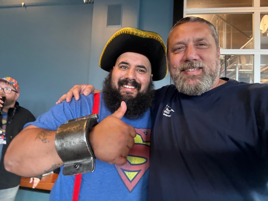 Two men posing for a selfie, one wearing a pirate hat and a blue Superman shirt while giving a thumbs up, and the other man with a beard in a dark shirt, smiling.