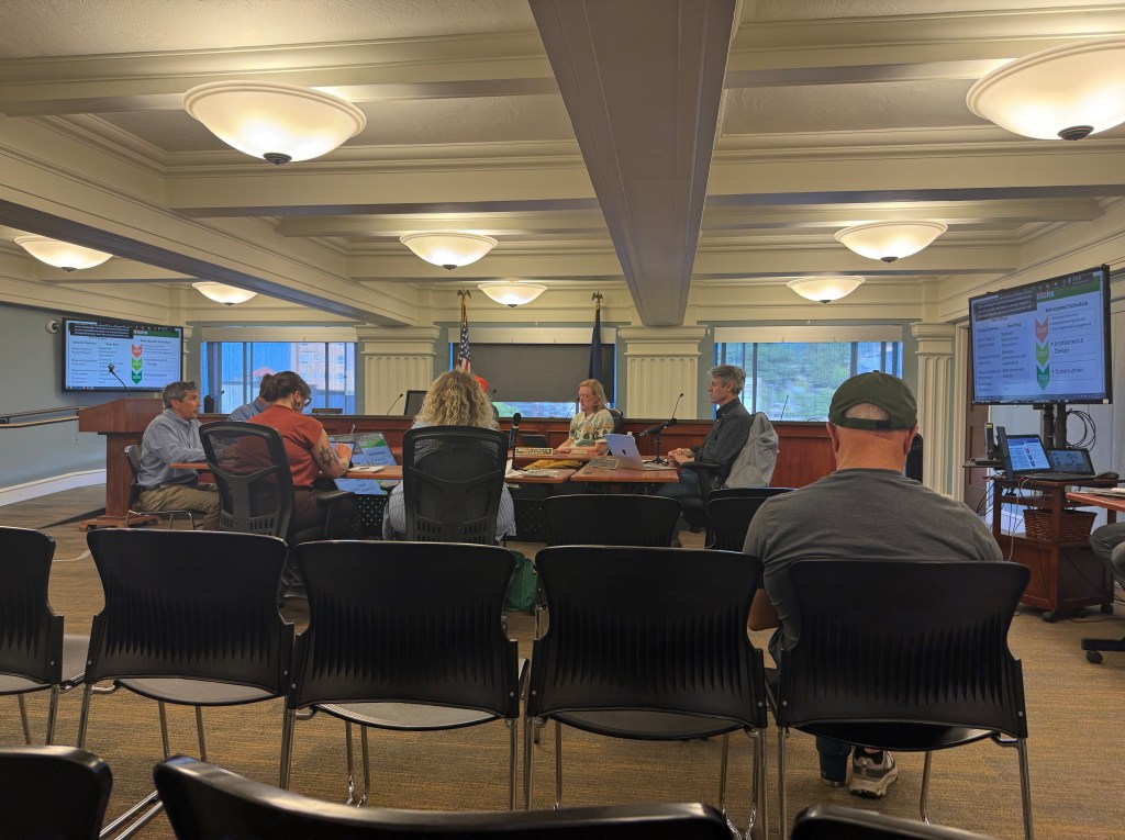 A meeting in a formal room with attendees seated and participating. Two large screens display information on a presentation, while a few individuals sit at a table discussing. The room features modern decor and ample lighting.