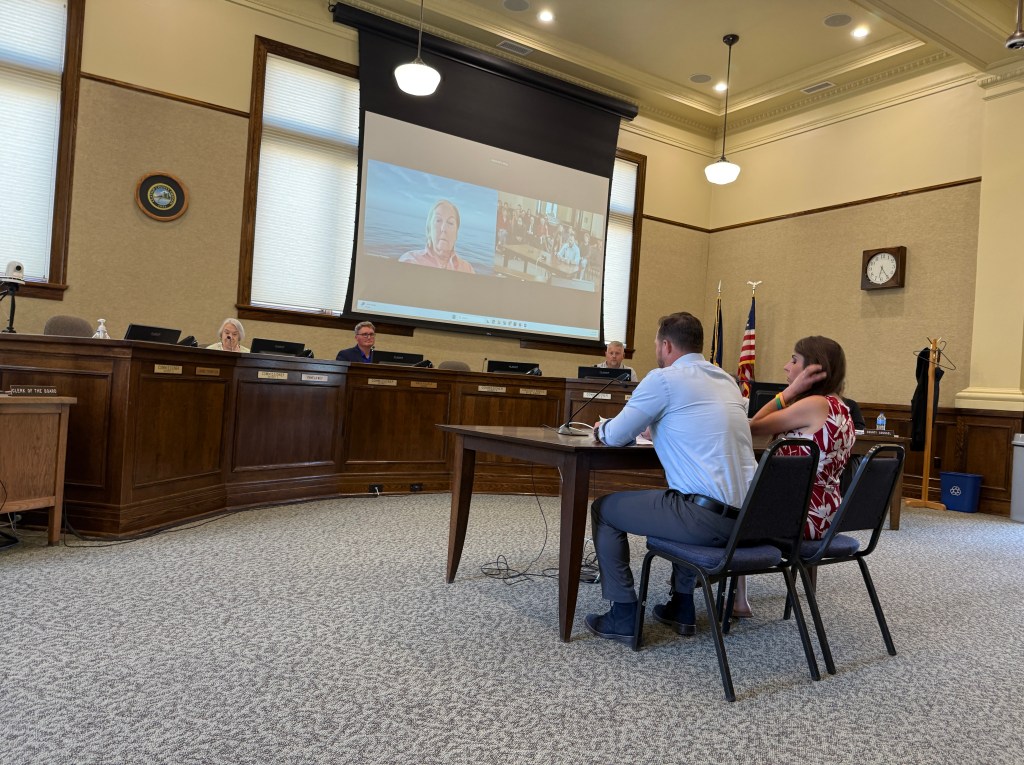 A municipal meeting in a chamber where two individuals are seated at a table addressing a panel of officials. One panel member is visible on a video screen in the background. There are flags and various documents present in the room.