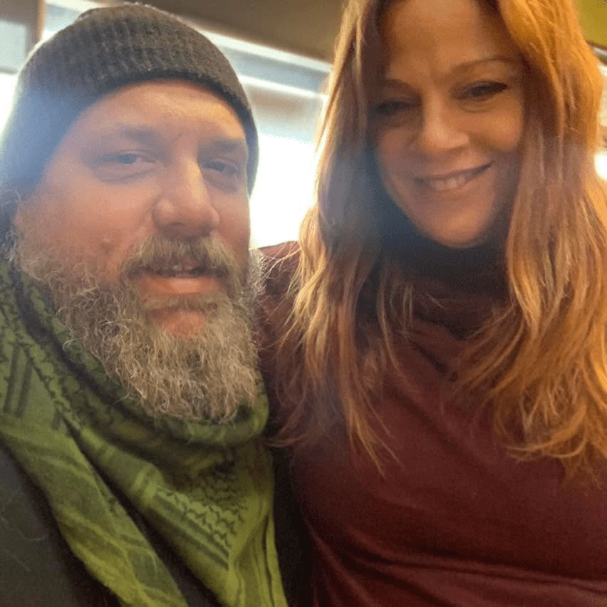 A smiling man with a beard wearing a green scarf and a black beanie poses for a selfie with a woman who has long red hair, both appearing happy and friendly.
