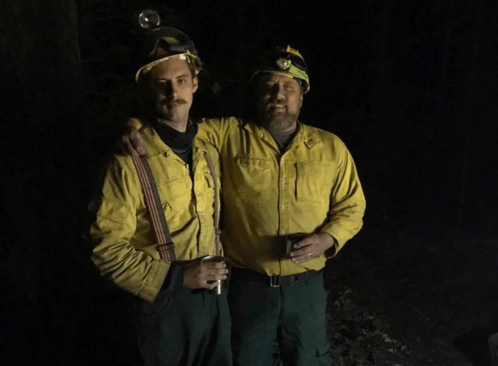 Two firefighters posing together in a forested area at night, wearing yellow firefighting gear and headlamps.