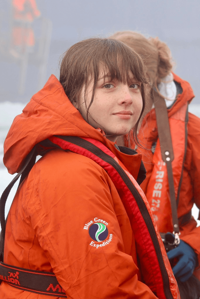 A young person wearing an orange waterproof jacket with a logo on the sleeve, looking towards the camera with a neutral expression.