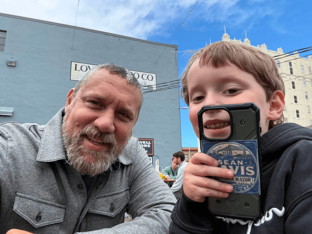 A smiling man and a young boy pose together, with the boy holding a phone case displaying a design. They are outdoors, with a blue sky and a building in the background.
