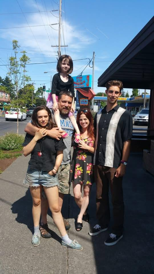 A family portrait outdoors on a sunny day, featuring five individuals: a man holding a young girl on his shoulders, a woman hugging him, another woman beside them, and a young man standing to the right.