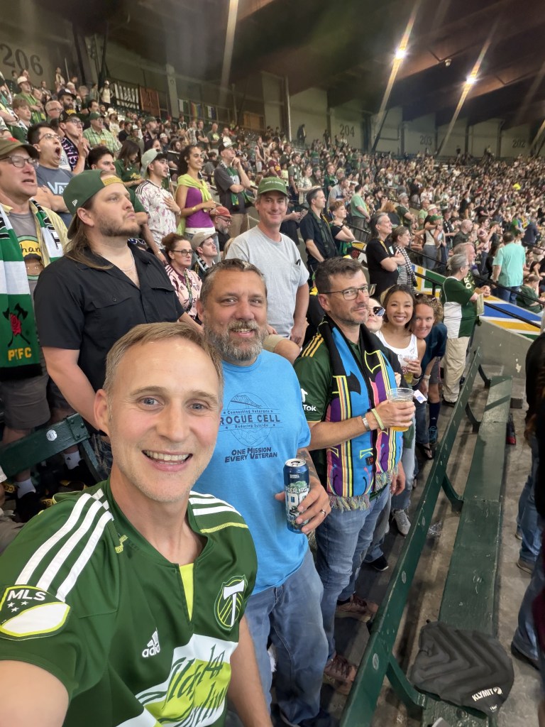 A group of enthusiastic fans at a soccer game, smiling and holding drinks, wearing team jerseys and scarves, with a packed stadium in the background.