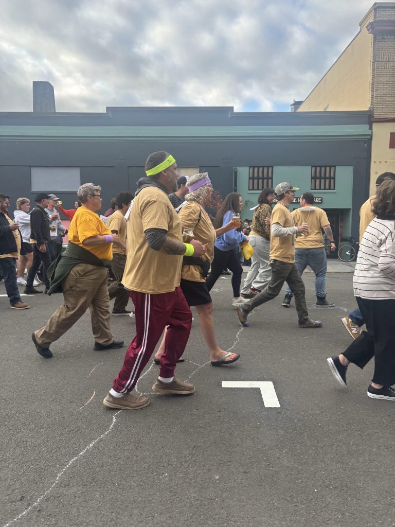 A diverse group of people in yellow shirts marching down a street, some wearing headbands and casual pants, with cloudy skies above.