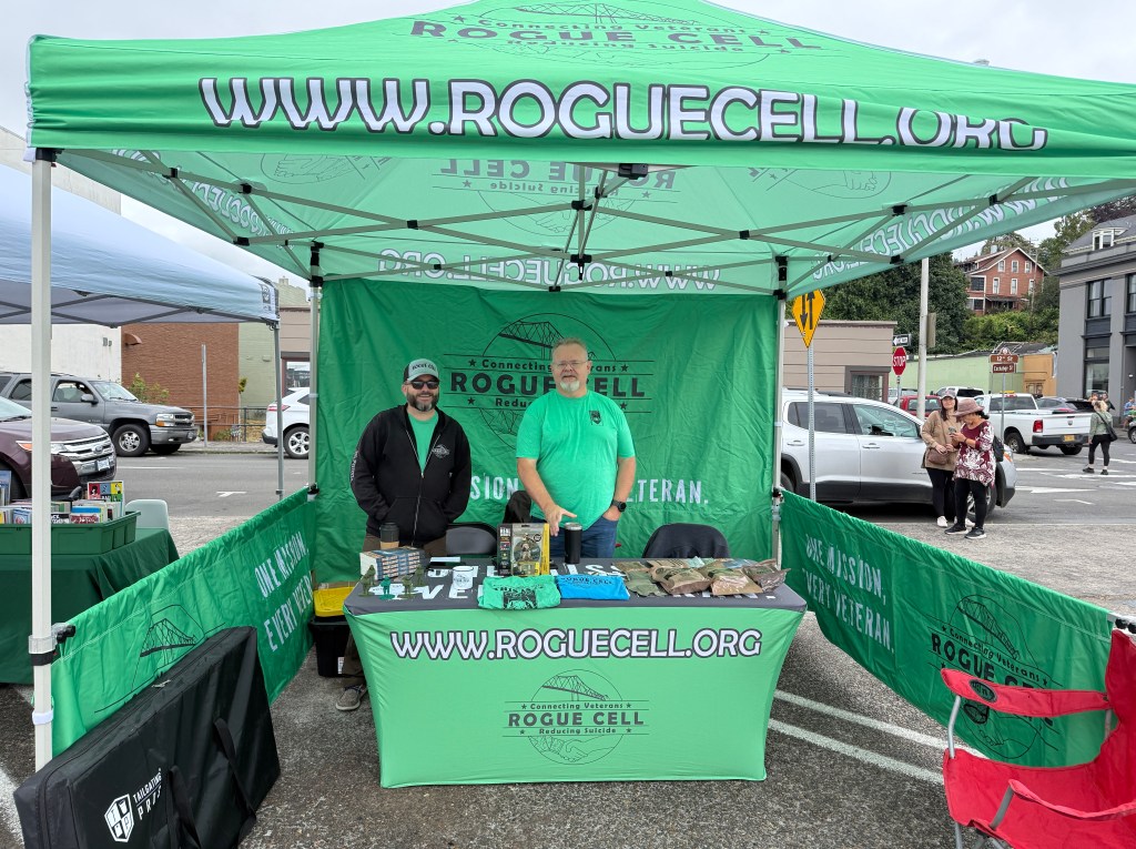 A booth set up for Rogue Cell, featuring two individuals in green shirts, surrounded by promotional materials. The tent displays the website www.roguercell.org and has signs about supporting veterans.