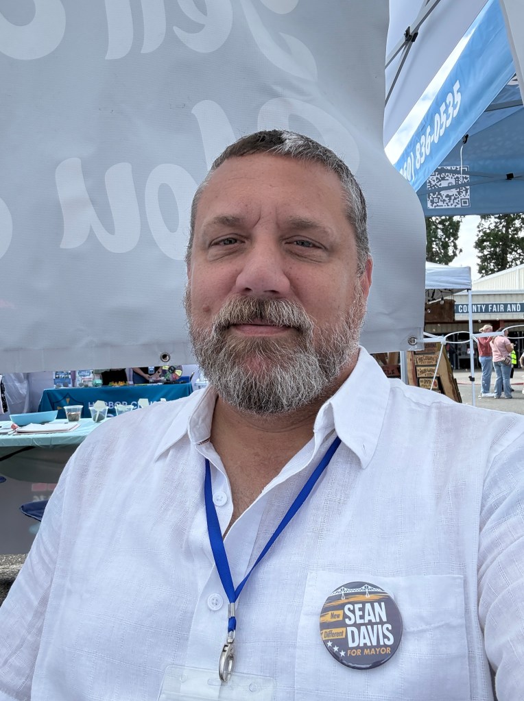 A man with a beard wearing a white button-up shirt and a blue lanyard, sitting at a table with a button that reads 'Sean Davis for Mayor' against a backdrop of banners at an outdoor event.
