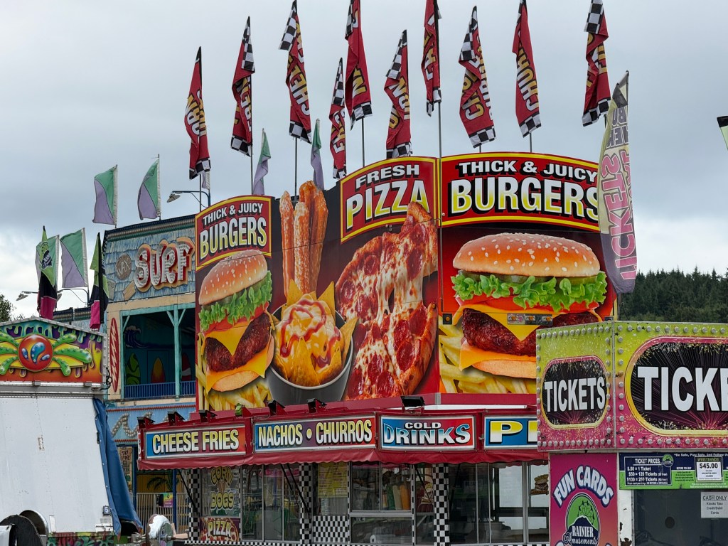 Colorful signboard featuring various food options like burgers, pizza, nachos, and drinks at a fair or festival food stand.