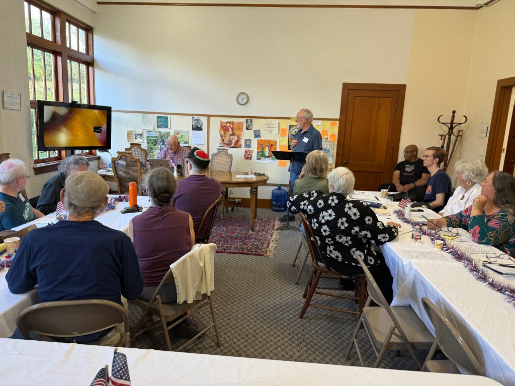 A group of people sitting at a table in a meeting room, engaged in a discussion. One person is giving a presentation at the front, with a display screen showing an image. The room has large windows and various posters on the walls.