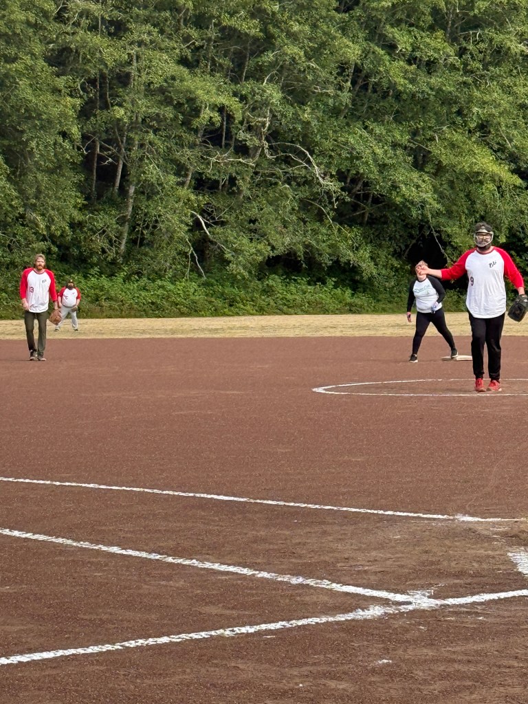 A group of four people playing softball on a dirt field surrounded by trees.