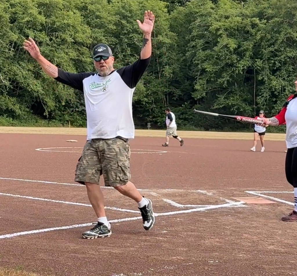 A man with raised arms celebrating on a softball field, wearing a white and black shirt with camo shorts. Other players are visible in the background.