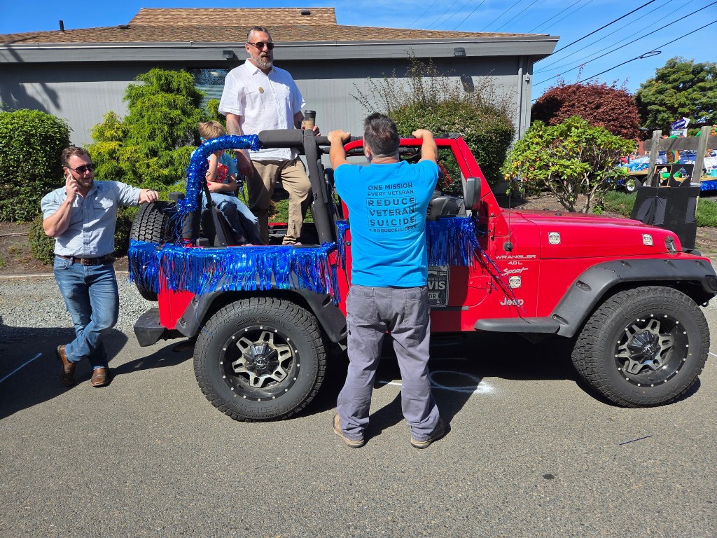 A red Jeep Wrangler decorated with blue fringe, parked on a street. Two men are standing near the vehicle, one on the phone and another wearing a blue shirt with a message about reducing veteran suicide. A child is seated in the Jeep, and a house and greenery are visible in the background.