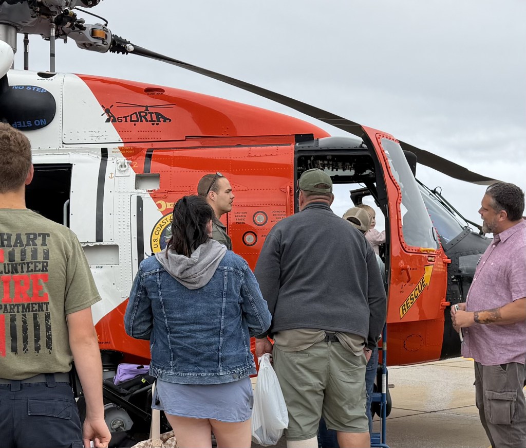 A group of people gathered around a red and white helicopter, with one person entering the cabin. The helicopter features U.S. Coast Guard branding and is stationed on a tarmac.