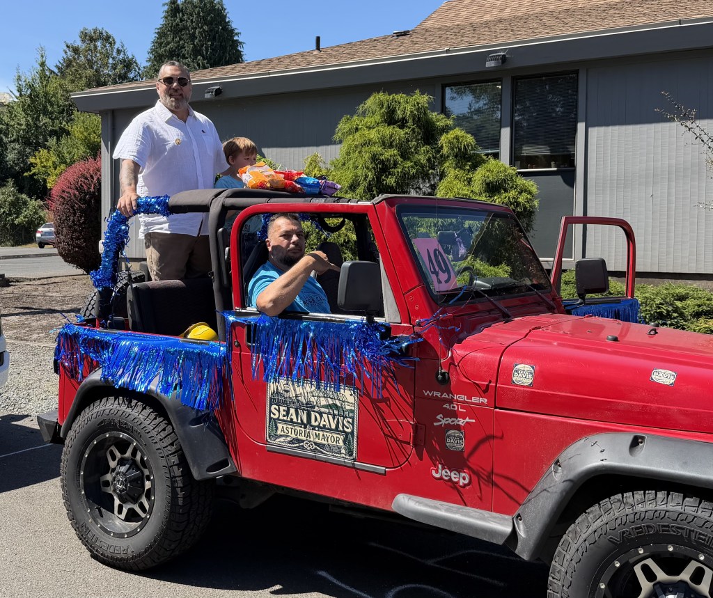 A decorated red Jeep with blue tinsel, featuring two men and a child. One man is driving while another stands beside the vehicle, showcasing a 'Sean Davis for Mayor' sign.