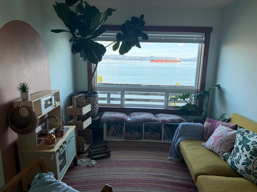A cozy living room with a yellow couch, a small play kitchen, potted plants, and a view of the water and a cargo ship through a large window.