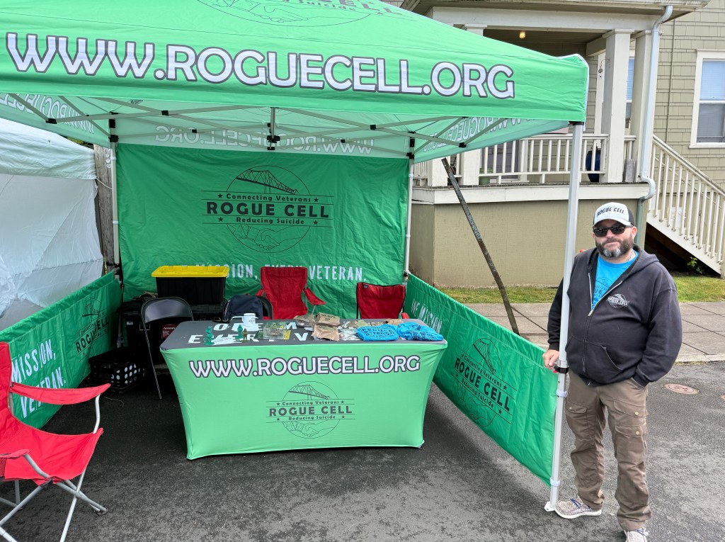 A booth set up for Rogue Cell with a green canopy displaying the organization’s name and slogan about reducing veteran suicide. A man stands next to the booth, which features tables with information and merchandise.