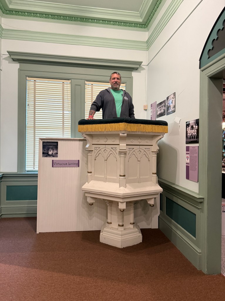 A man stands on a decorative pulpit in a museum setting, with green trim and large windows in the background. A sign nearby reads 'Virtuous Living'.