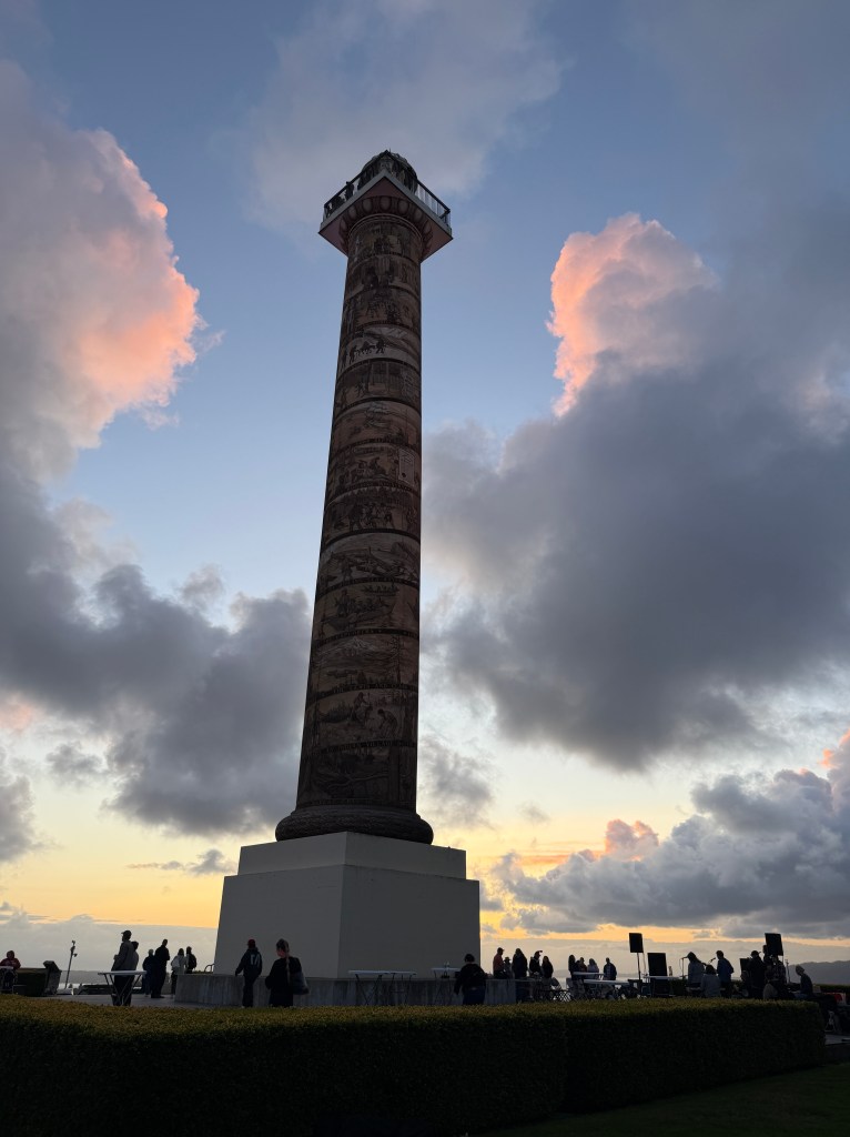 A tall decorative column featuring intricate carvings, set against a backdrop of pastel clouds during sunset, with people gathered around its base.