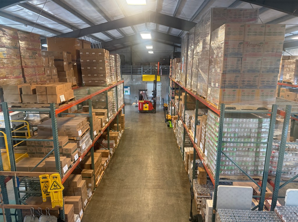 Interior view of a large warehouse with high shelves filled with stacked boxes and pallets, showcasing organized storage and inventory management.