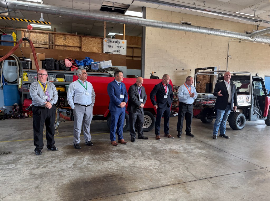 A group of seven men in business attire stands in a fire department garage, with equipment and a red vehicle in the background. One man is speaking while others listen attentively.