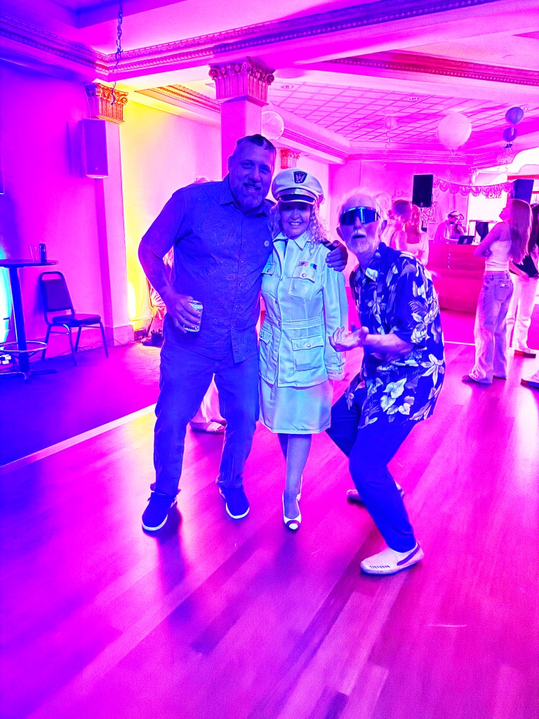 Three people pose for a photo in a colorful party setting, with vibrant purple lighting. One person is wearing a police-style outfit, while the others are dressed casually; all appear to be enjoying the event.
